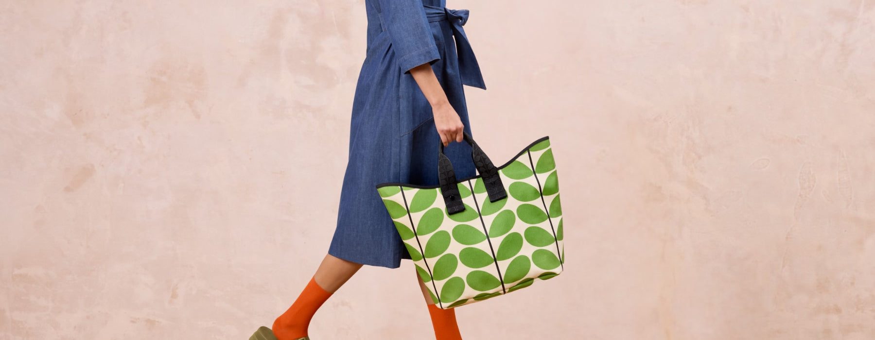 A large tote bag featuring a green stem print, being carried by grab handles by a women in a blue dress