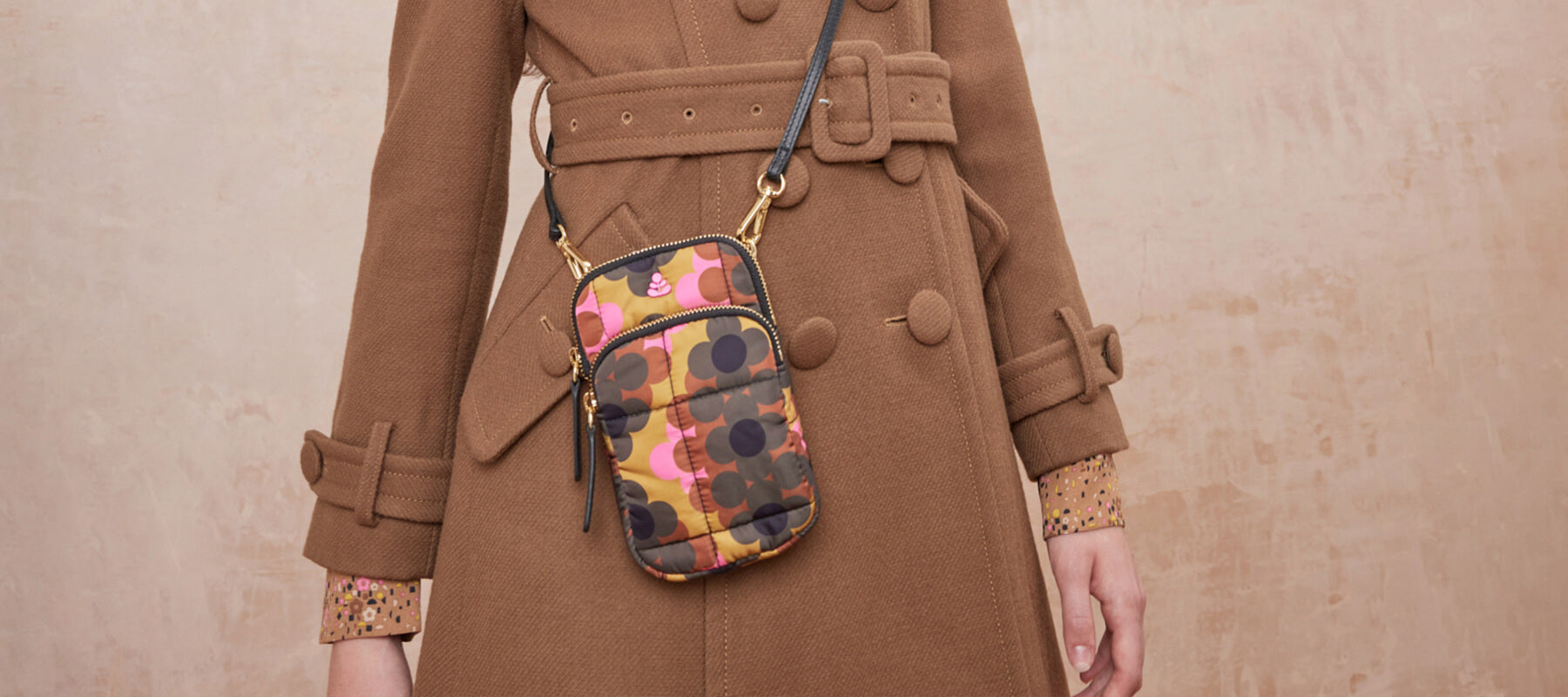 A women wears an Orla Kiely crossbody phone pouch with a retro inspired pattern in brown, pink and yellow.