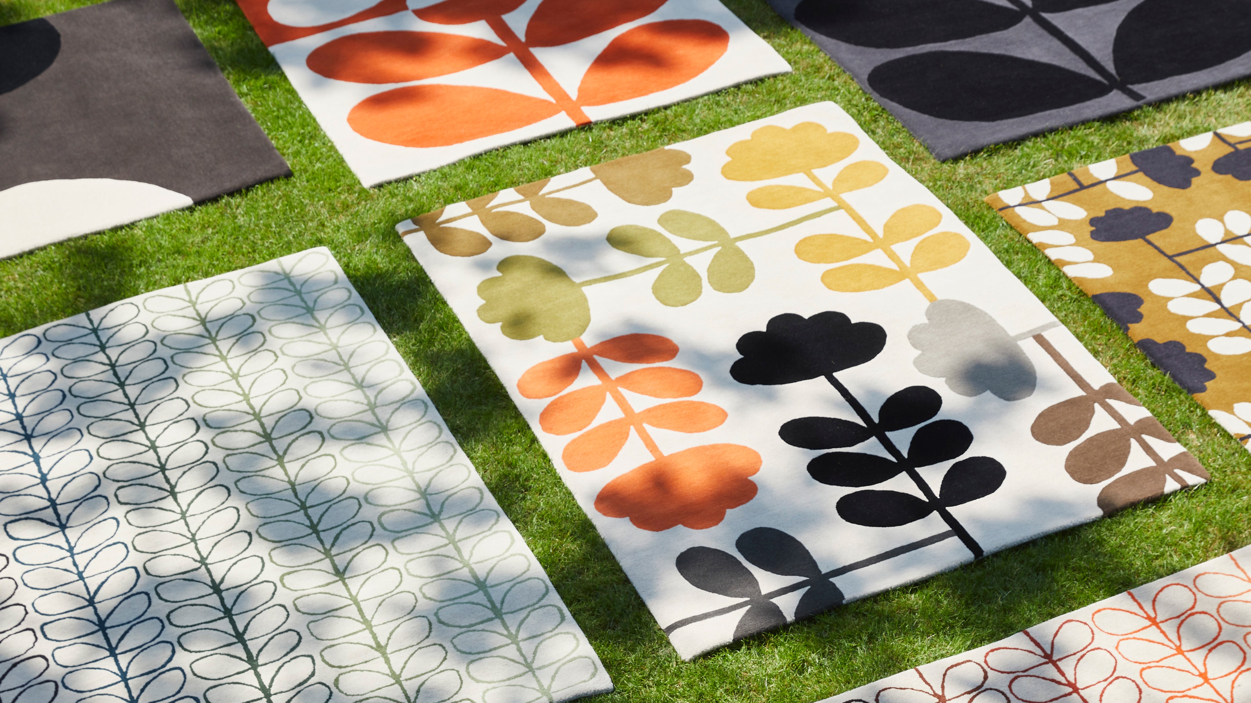 A number of multi coloured patterned rugs lie together on a floor in retro colours of orange, green and brown.