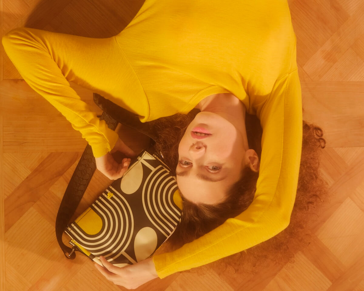 Model in yellow lying on the floor with an Orla Kiely mini rounder bag 