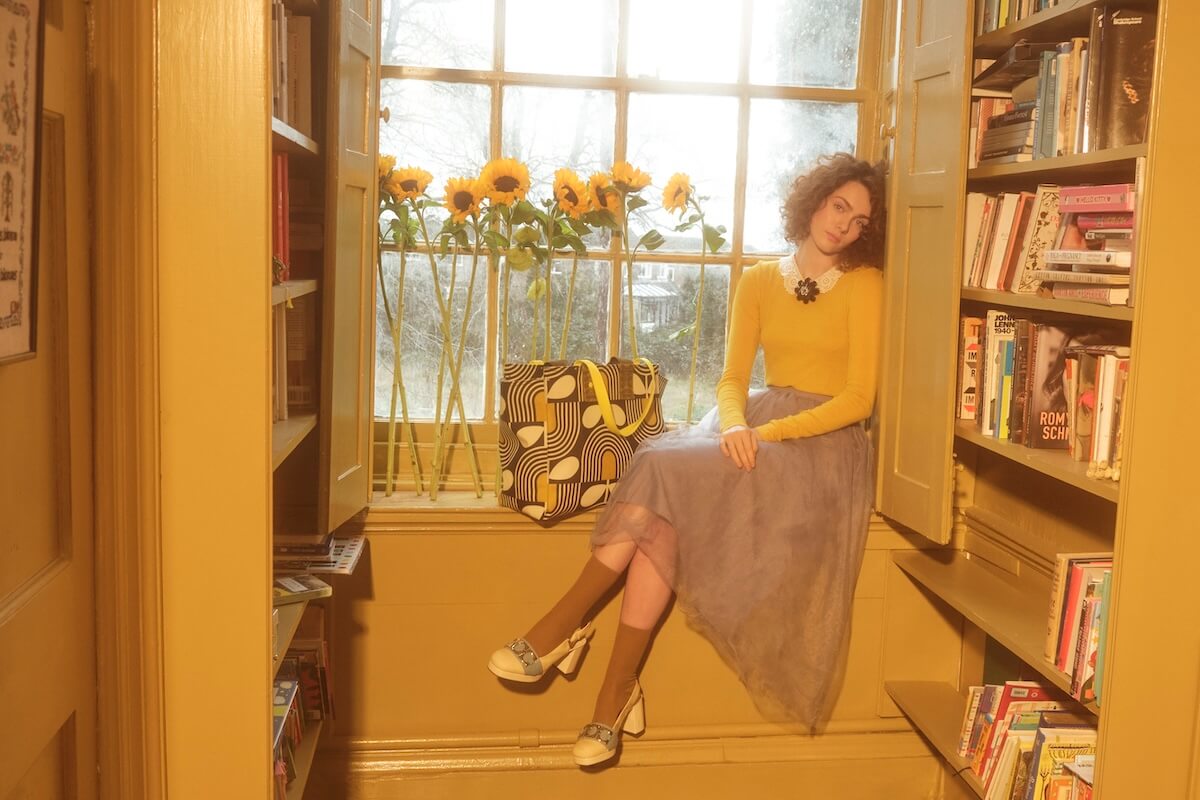 Model sitting by book shelf next to Orla Kiely all in tote