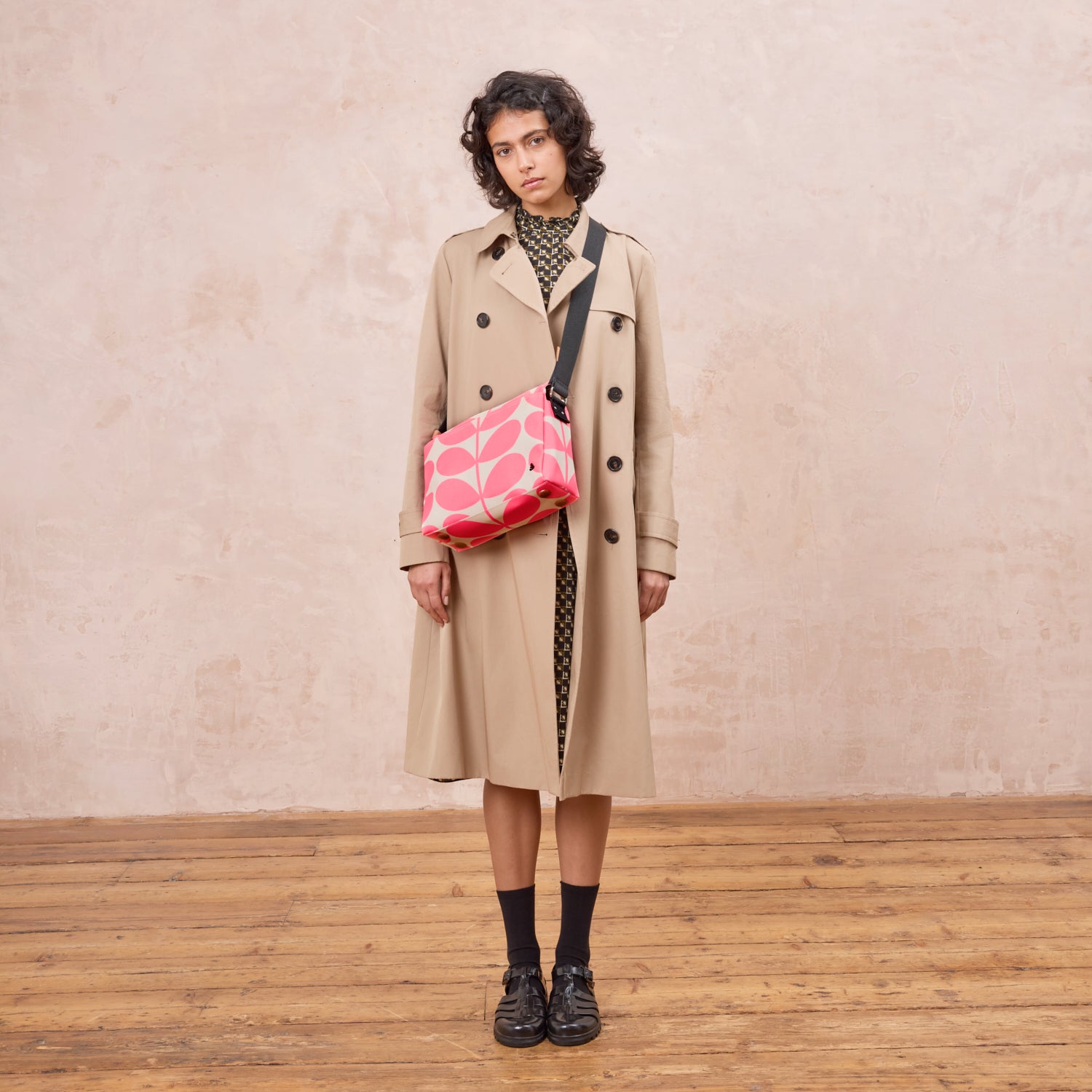 A woman in a beige trench coat wears the Fielder Crossbody bag with a neon pink steam print.