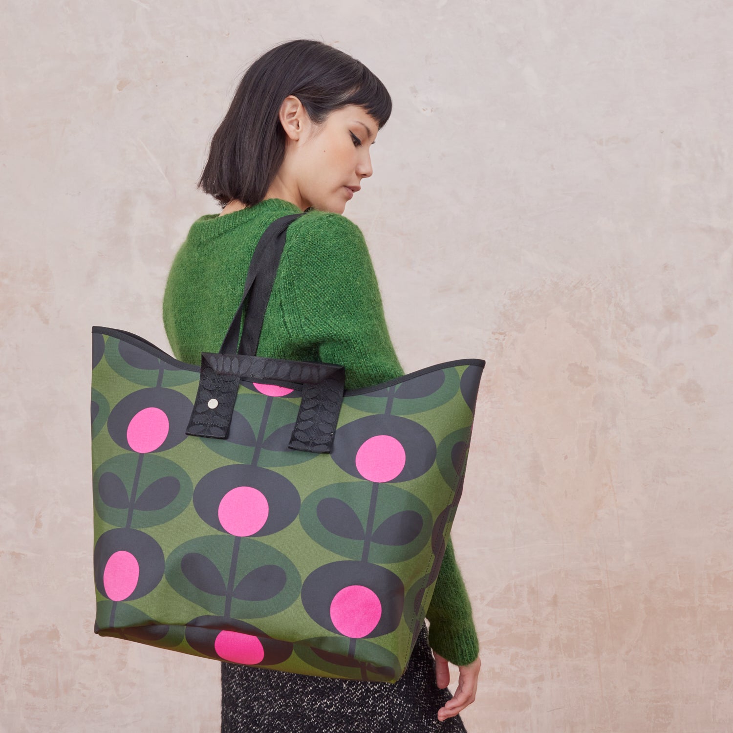 Woman carrying a large green tote bag with pink and black floral pattern with Orla Kiely webbing handles and shoulder strap.