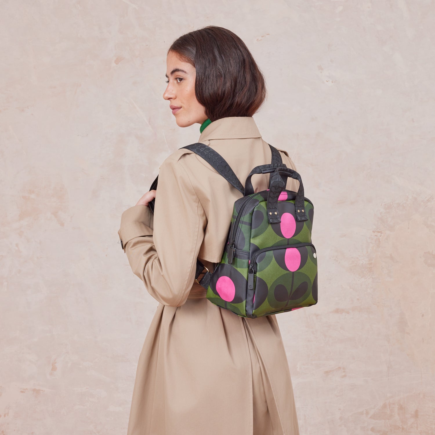 A womens wears the mini Lotta Backpack in a green stem print with bright pink dots, featuring a zipped front pocket, branded grab handles and adjustable shoulder straps.