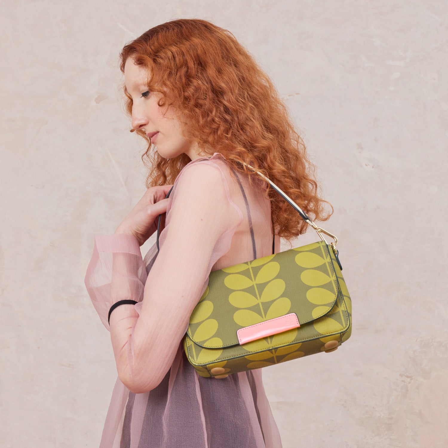 A woman wears a green stem patterned handbag with a pink branding over her shoulder