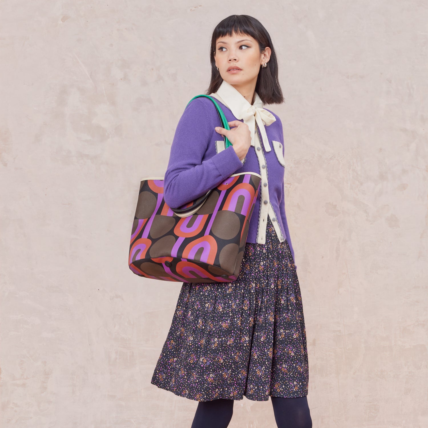 A woman wears a large tote bag over her shoulder that features purple, red, brown and black retro inspired pattern.