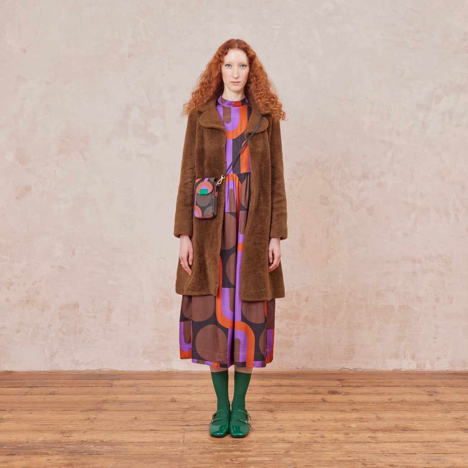 A woman wears a purple patterned phone pouch with a crossbody strap, she also wears a matching printed dress, brown coat and green shoes.