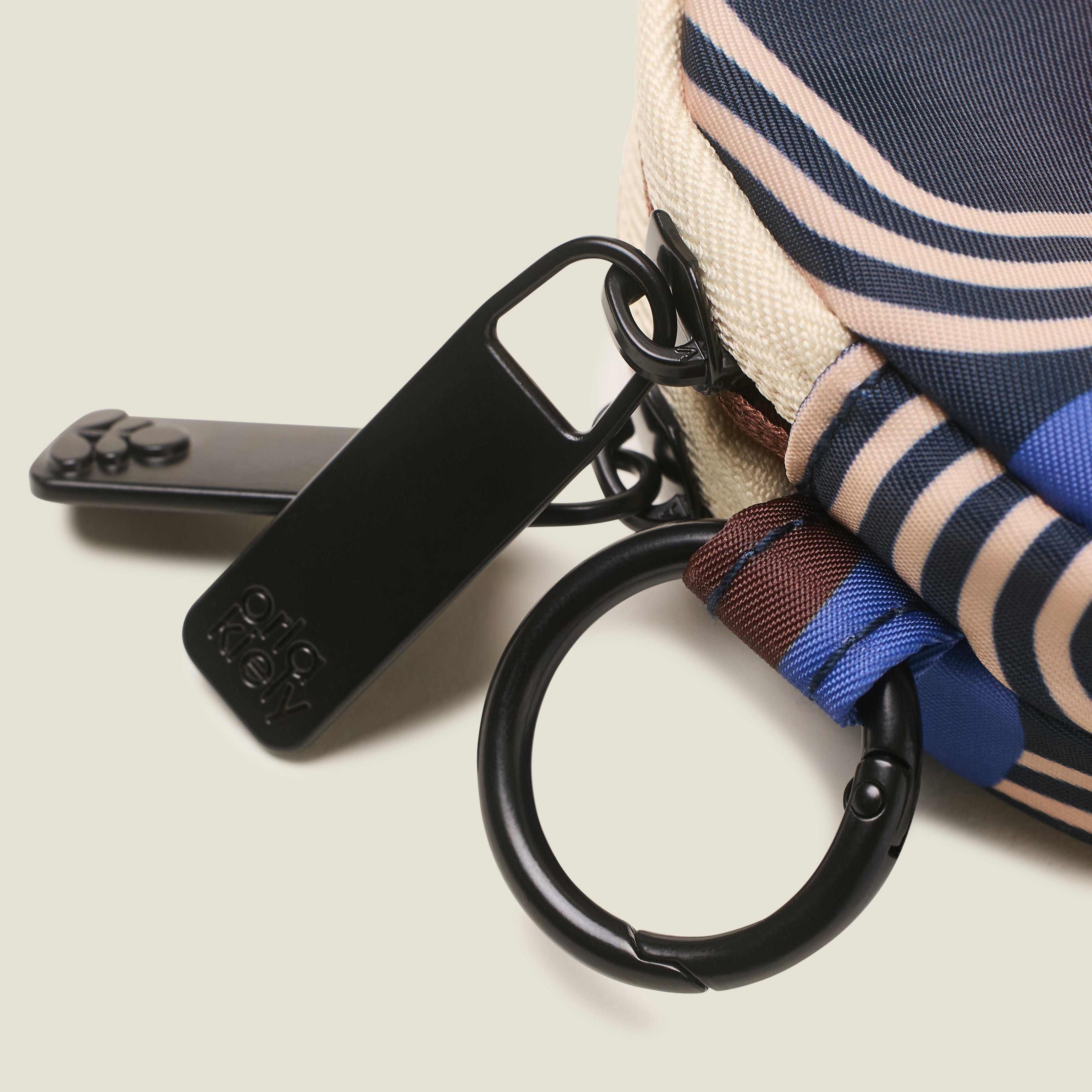 Close up of round coin purse, with a branded zip pulls in a blue, navy, brown and cream retro print with a ring clip on a beige background.