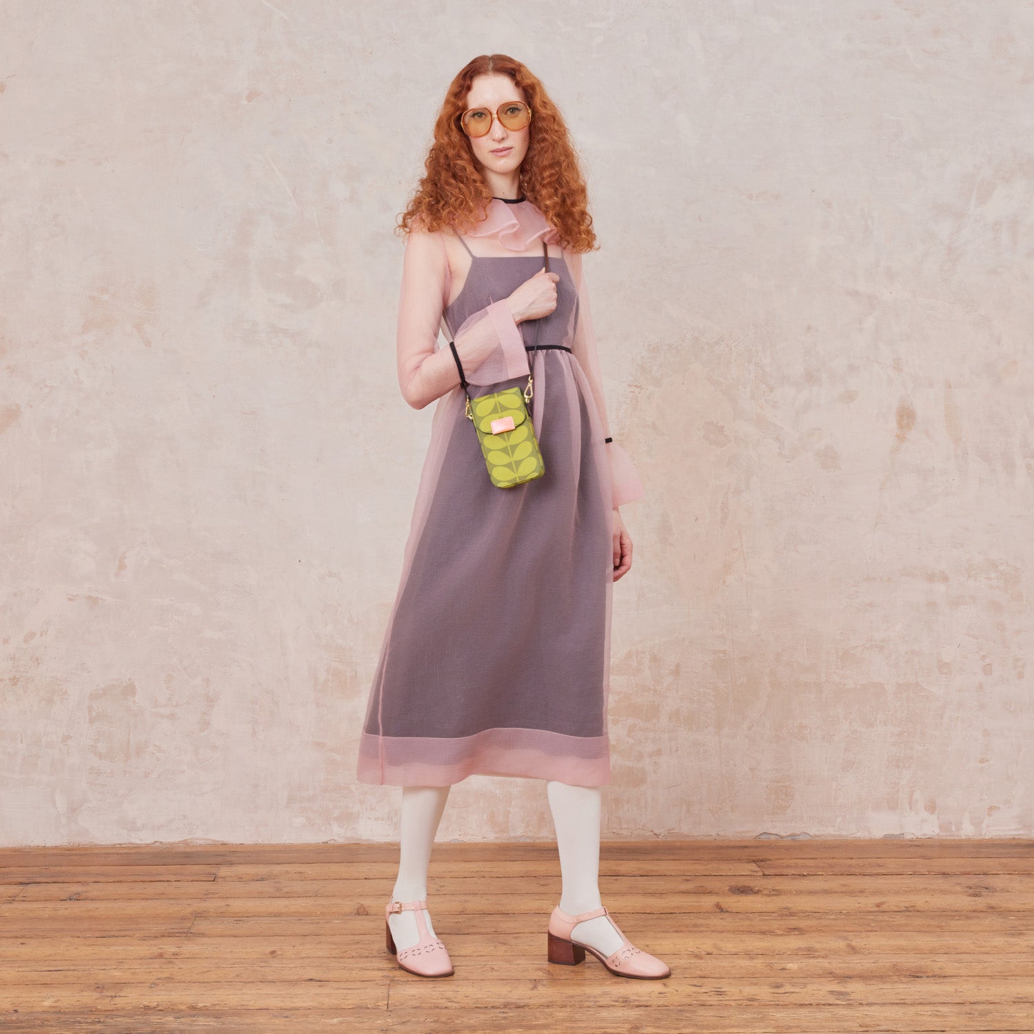 A woman in a pink sheer dress wears a green stem print phone pouch with leather crossbody strap and pink branded trims.