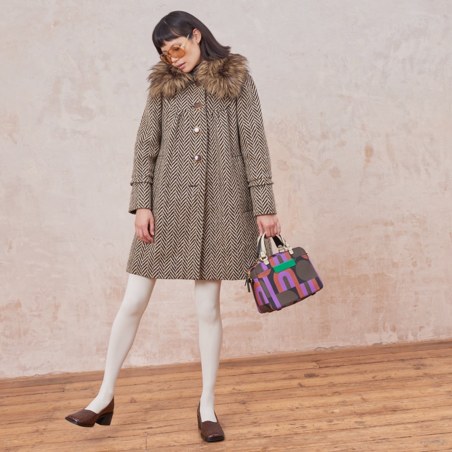 A woman holds the medium sized square handbag in a retro purple, red and brown print, featuring grab handles and zip closure.