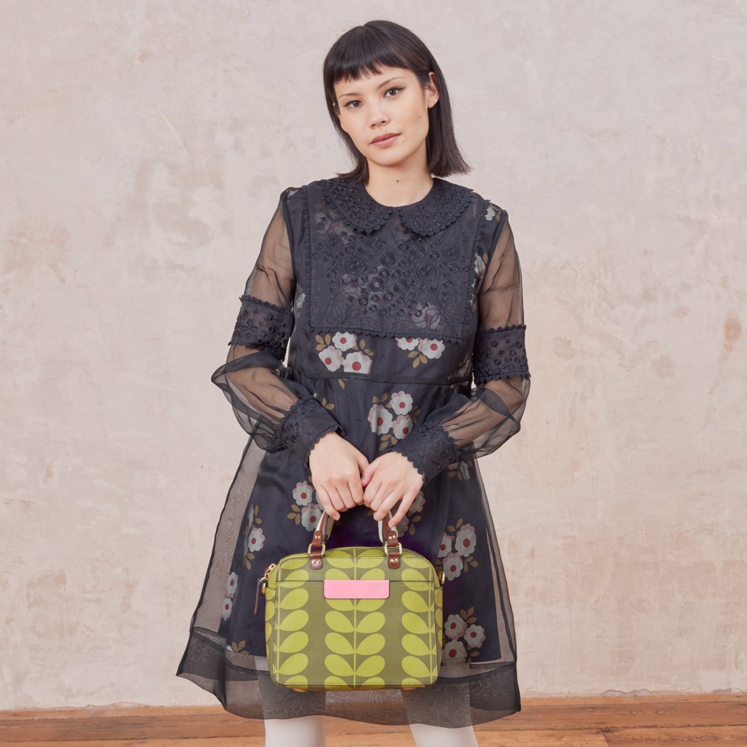 A woman holds a medium square handbag with green leaf pattern by two grab handles, with pink branding.