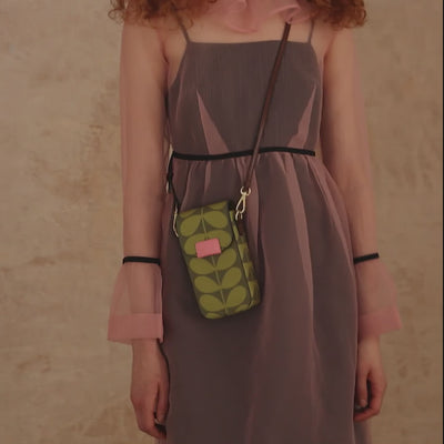 A woman in a pink sheer dress wears a green stem print phone pouch with leather crossbody strap and pink branded trims during a photo shoot.